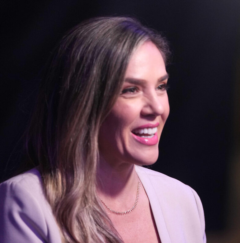 Close up headshot of Jaime Maggio wearing a beige blazer in front of a black background