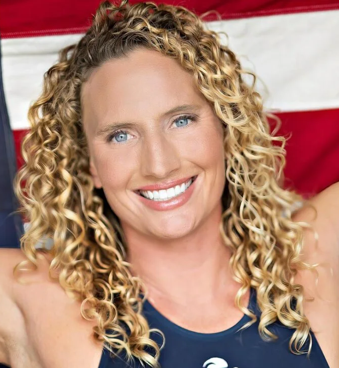 Professional headshot of Kaleigh Gilchrist wearing a navy swimsuit top posing in front of an American flag