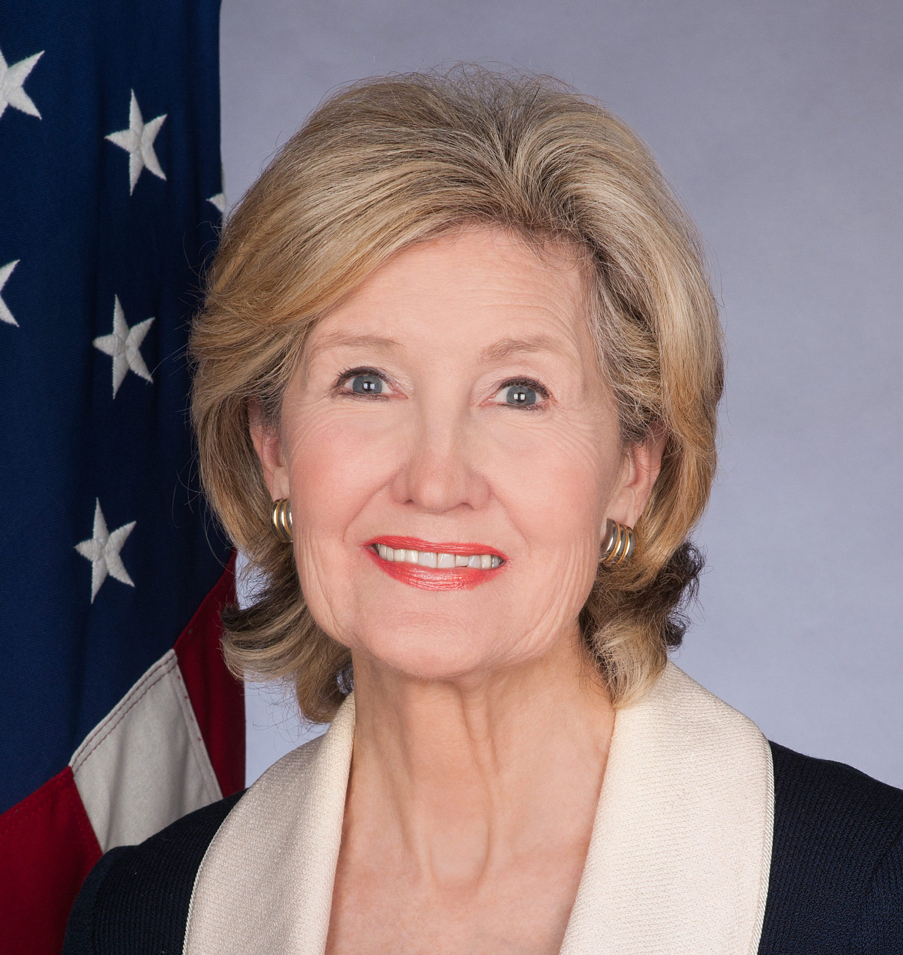 Professional headshot of Kay Bailey Hutchinson wearing a black blazer with a white collar posing in front of an American flag