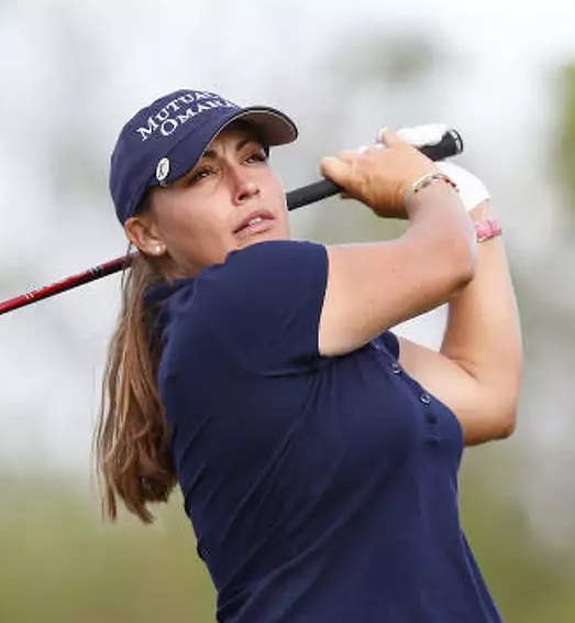 Action shot of Kelli Kuehne swinging a golf club outdoors wearing a navy blue shirt and navy hat