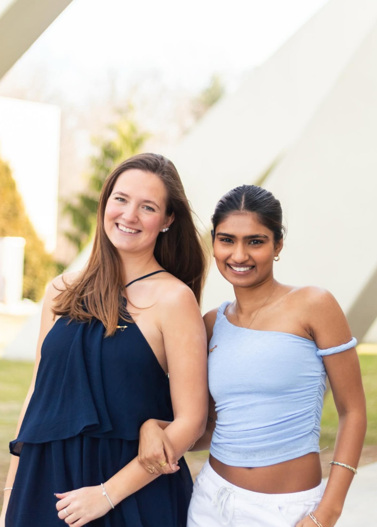 Two members wearing dark and light blue tank tops standing outdoors in front of a white building and green grass