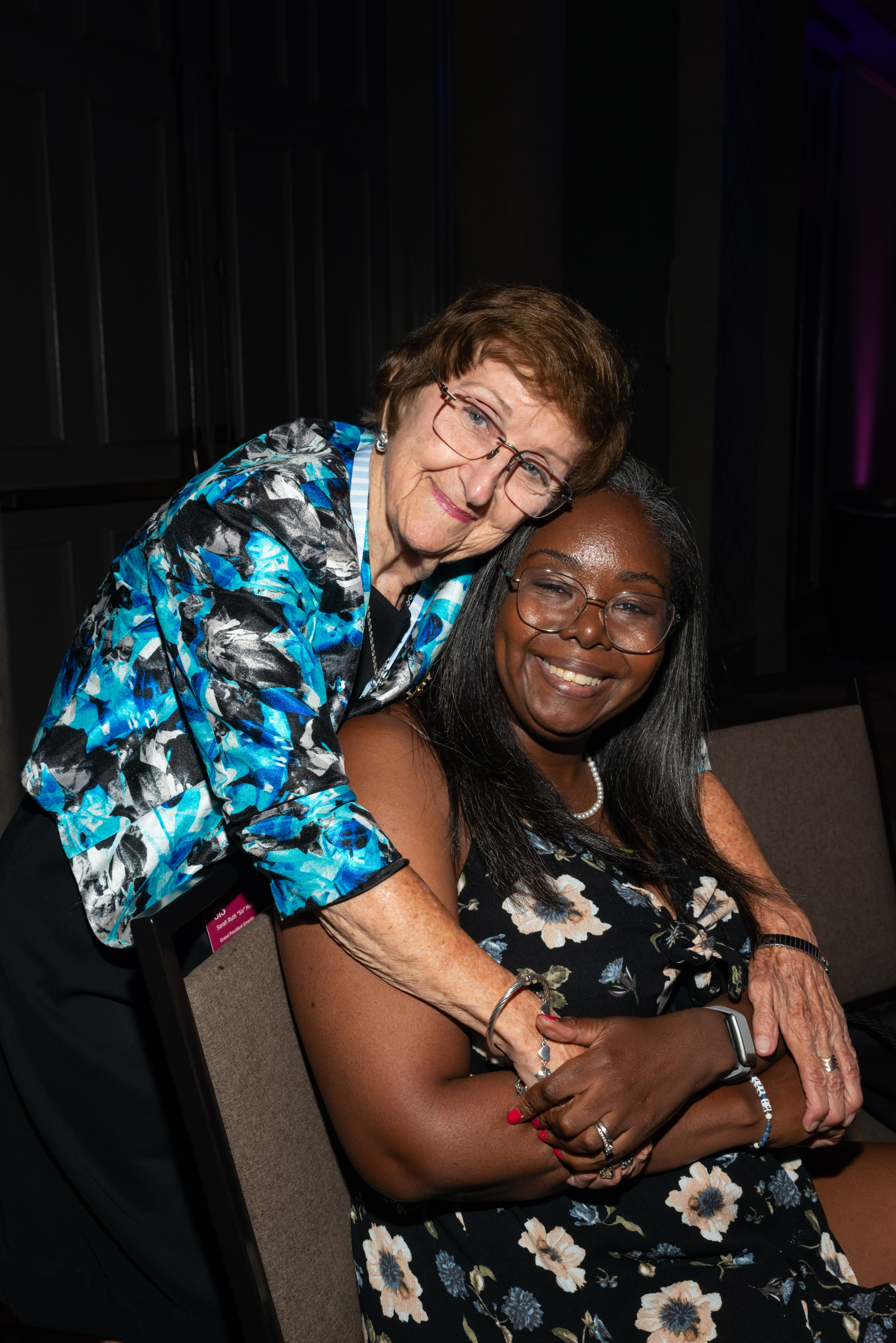 One alumnae member wearing a blue and black jacket leaning over a chair to hug a member wearing a black and yellow flowered dress