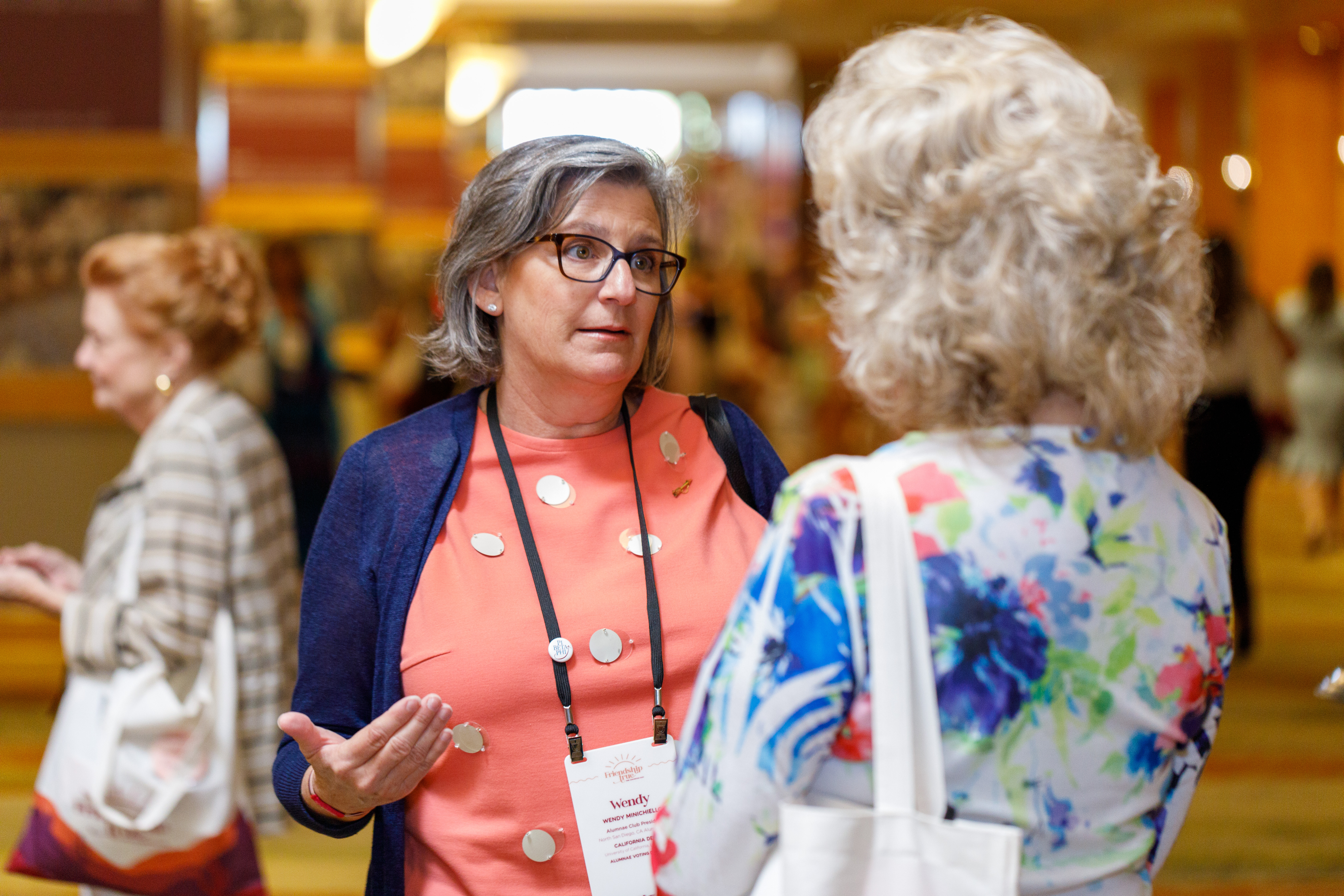 One alumnae member wearing an orange shirt speaking to an alumnae member wearing a blue and white flowered top with her back to the camera at a conference indoors with other members in the background