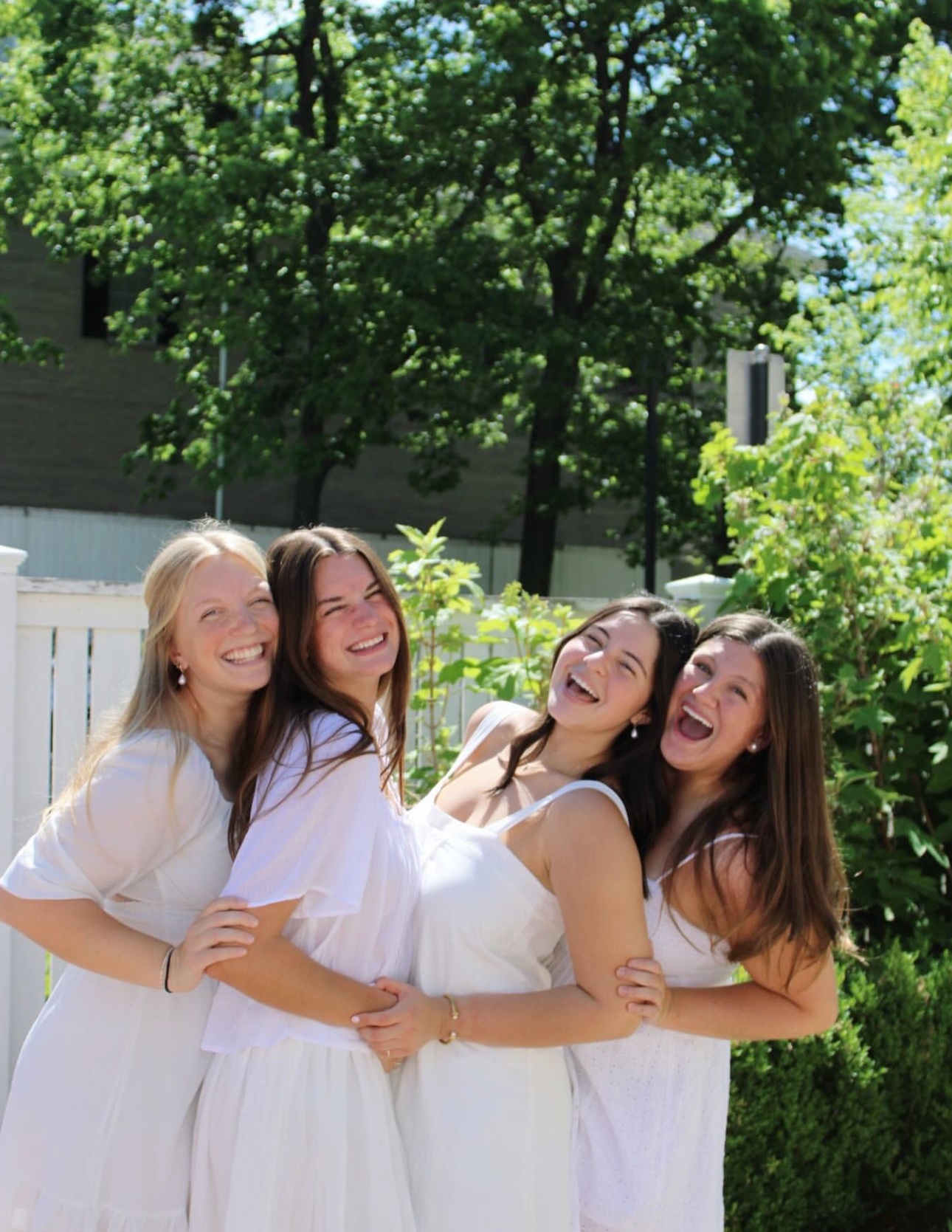 Four members wearing white dresses hugging outdoors in front of green trees