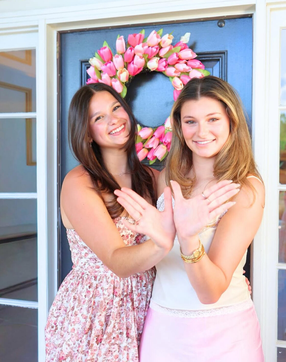 Two members wearing pink dresses standing outdoors in front of a dark door with a pink wreath holding their hands to make angel wings
