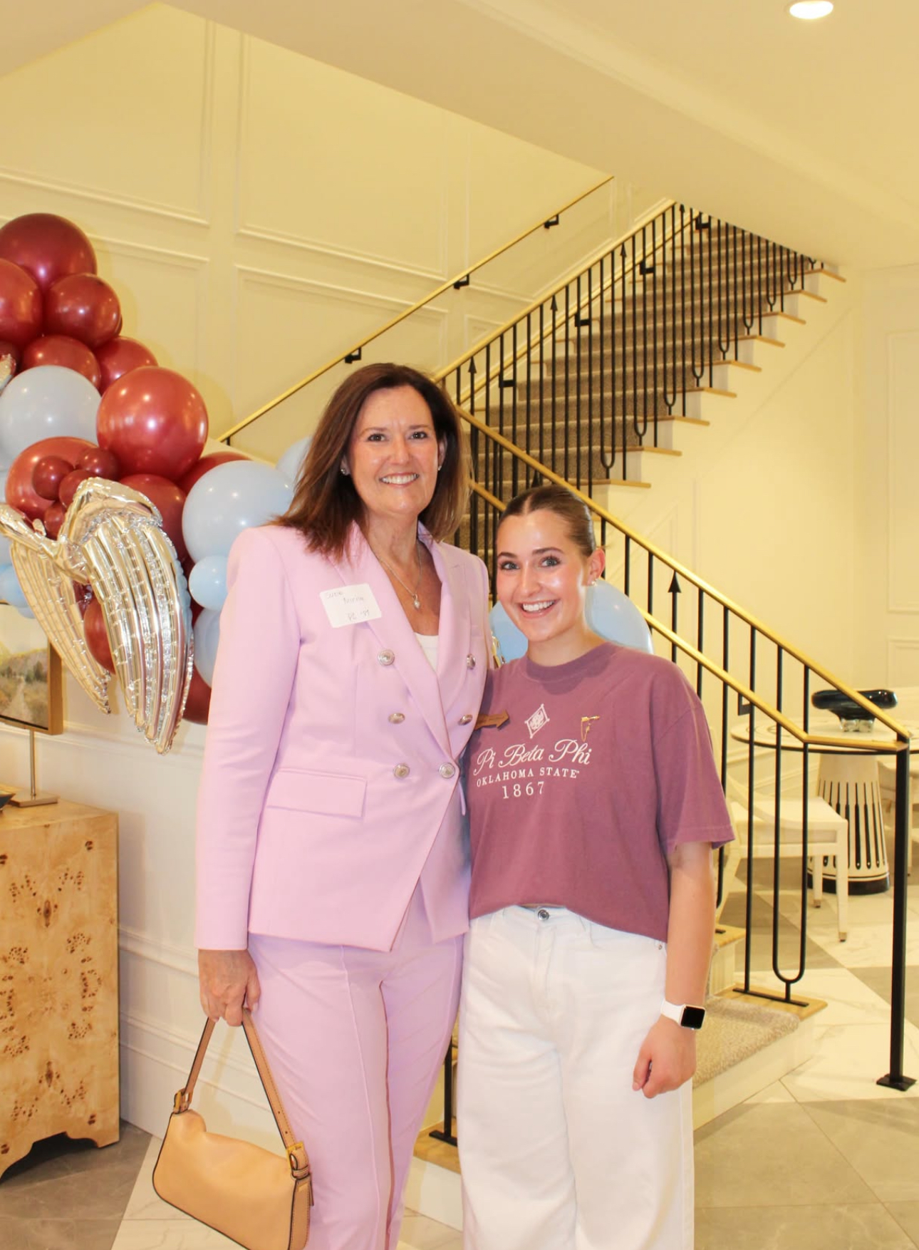 Two members standing indoors in front of a staircase and blue and red balloons wearing a light pink pantsuit and a dark pink t-shirt and white pants