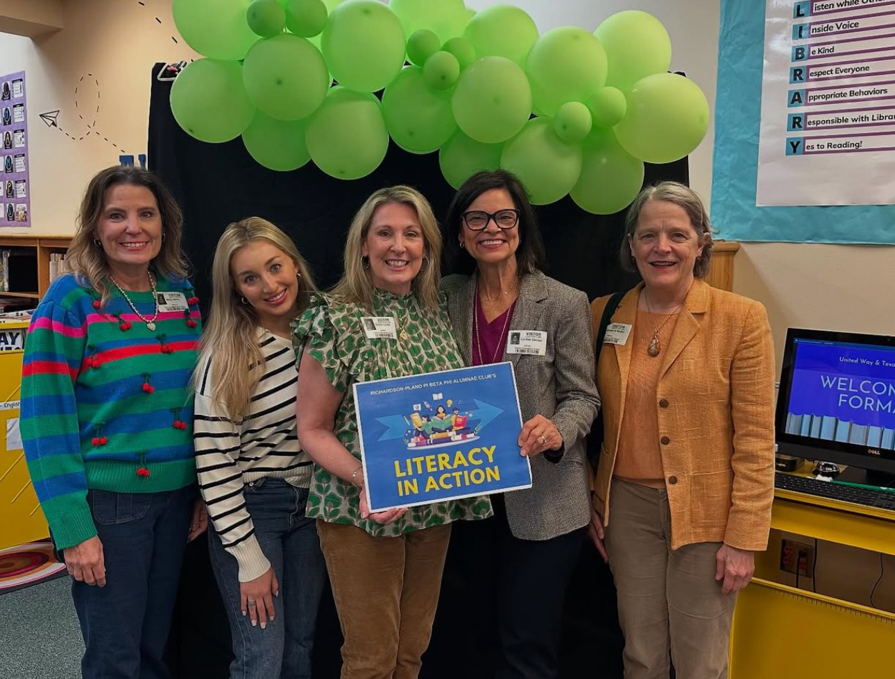 Five alumnae members holding a blue sign that reads, "Literacy in Action" standing in a classroom with a banner of green balloons