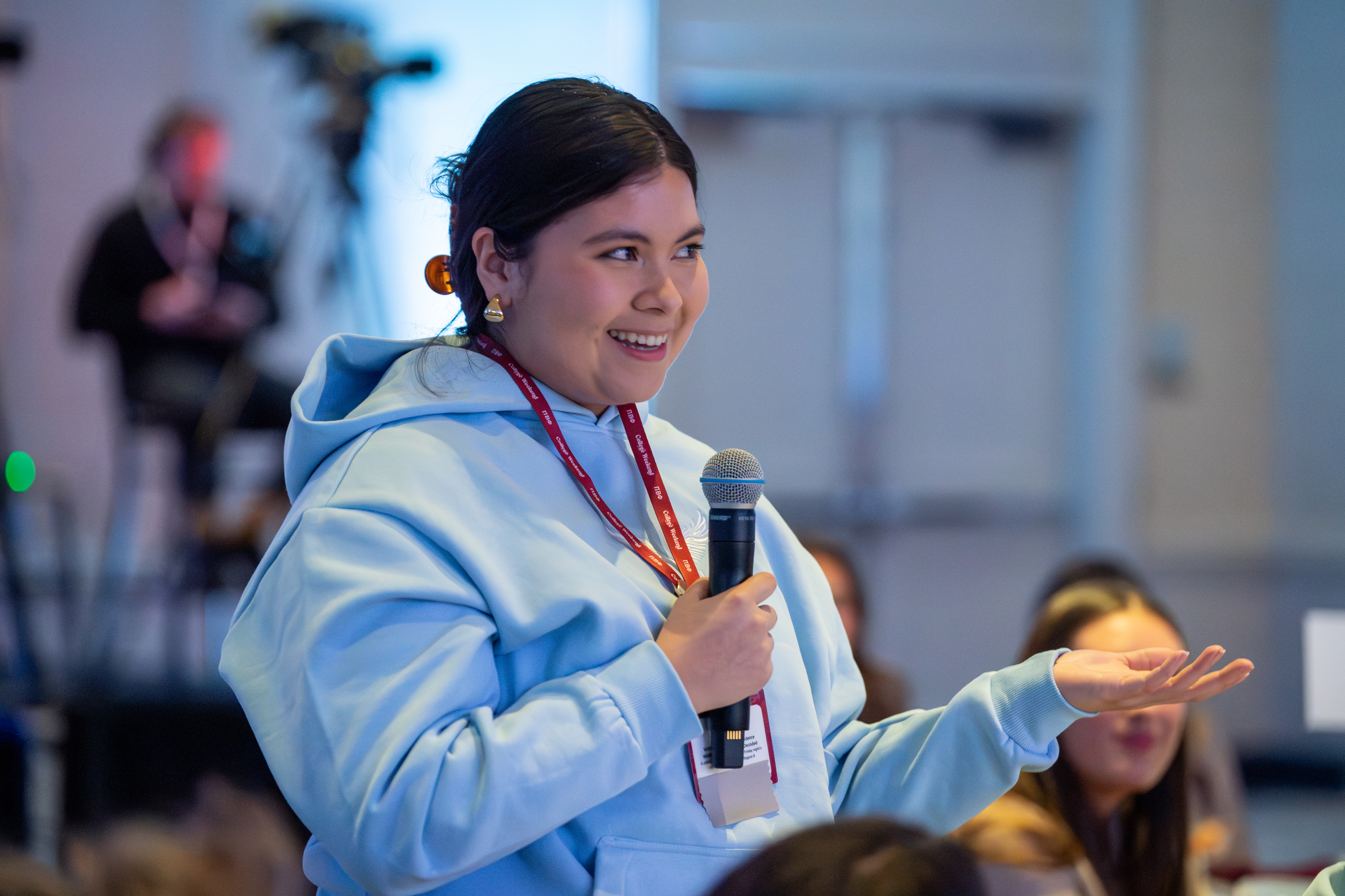 A member wearing a light blue hooded sweatshirt stands inside at a conference speaking into a microphone
