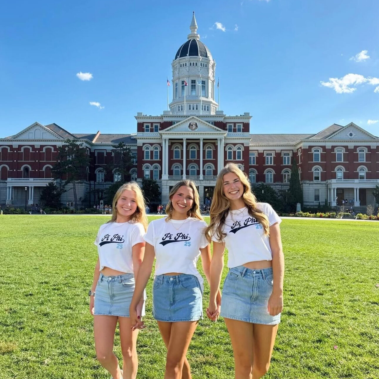 Three members holding hands wearings matching white shirts and denim skirts in front of a campus building