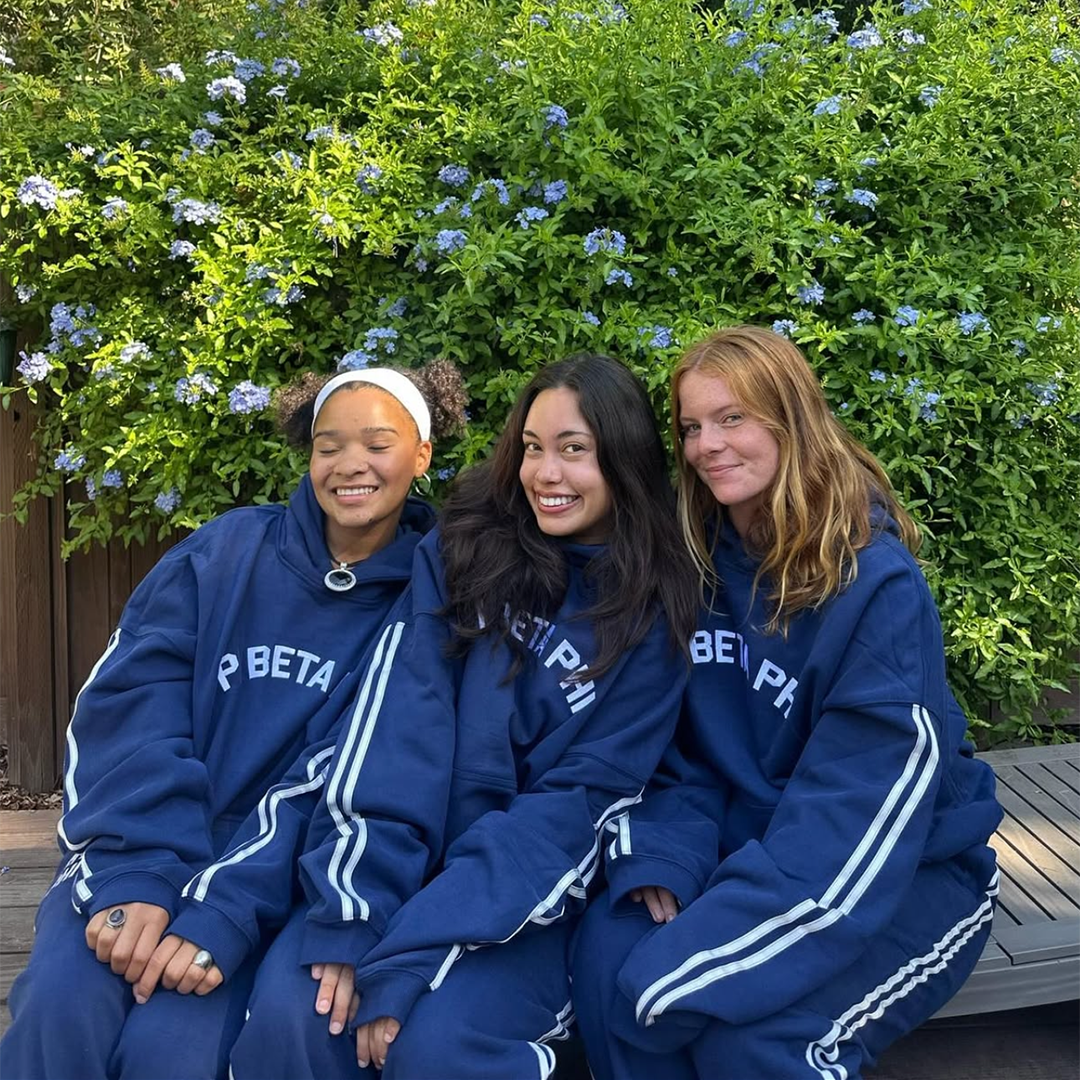 Three members wearing navy sweatsuits sitting in front of greenery