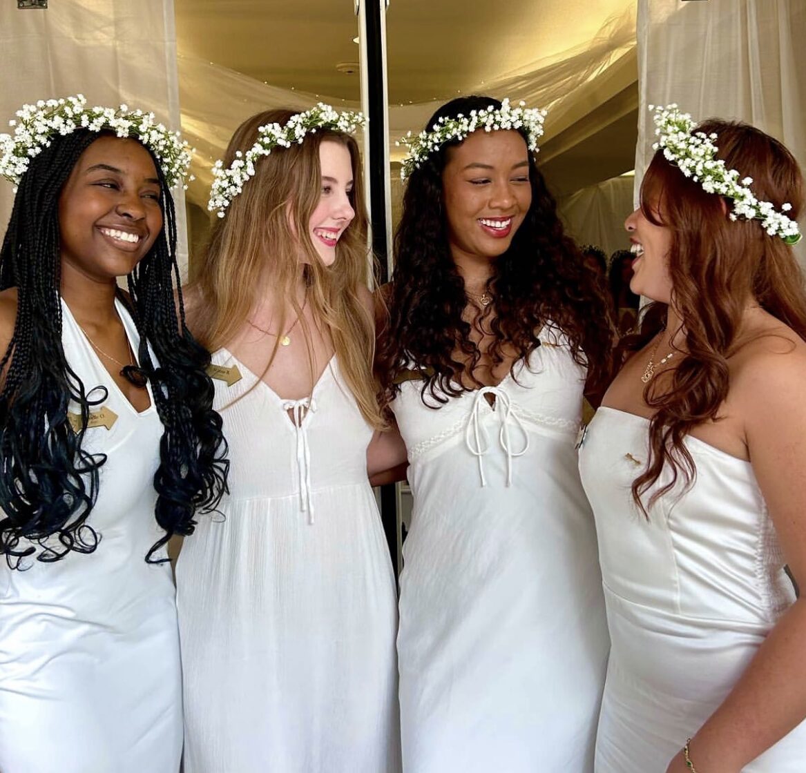 Four members wearing white dresses and white flower crowns smiling at one another