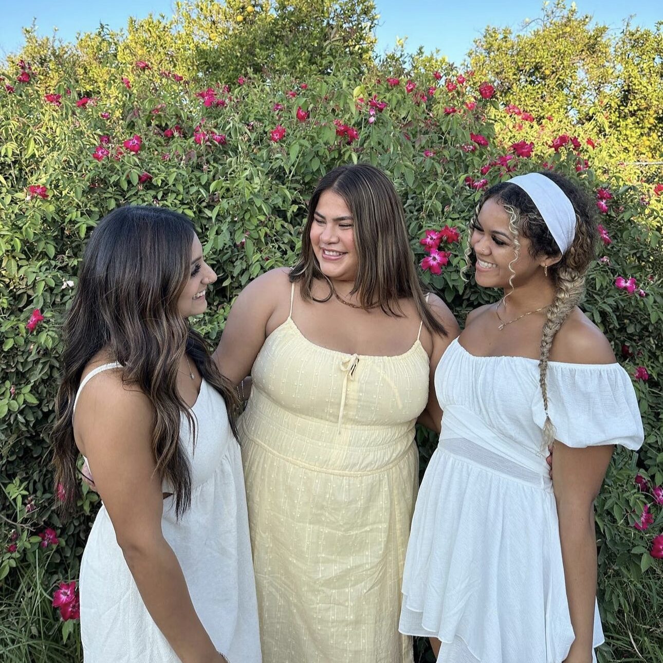 Three members smiling at each other in front of a green bush
