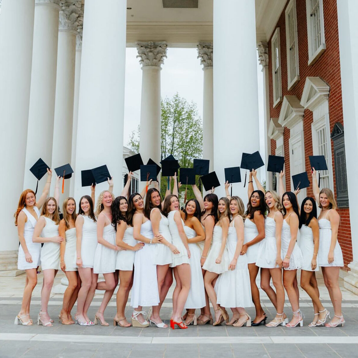 Several members wearing white and holding graduation caps in front of columns on a building