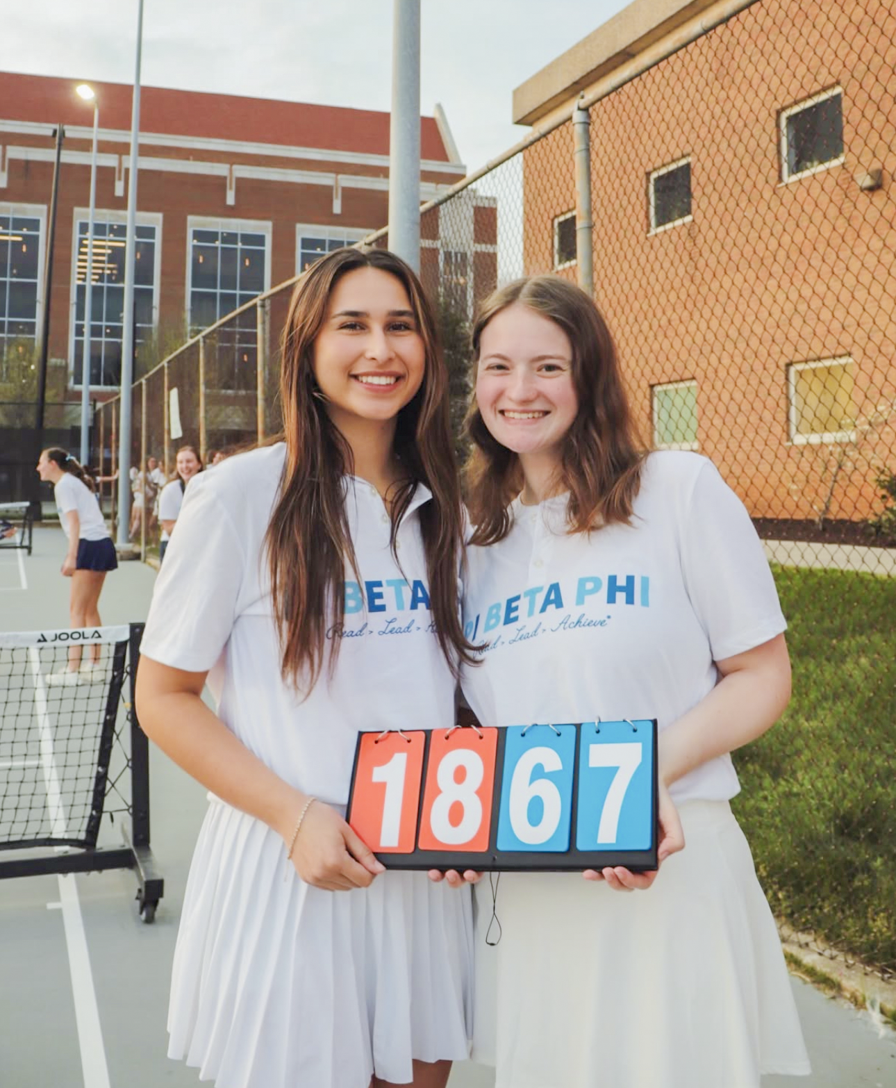 Two individuals standing outdoors holding a scorecard that reads 1867