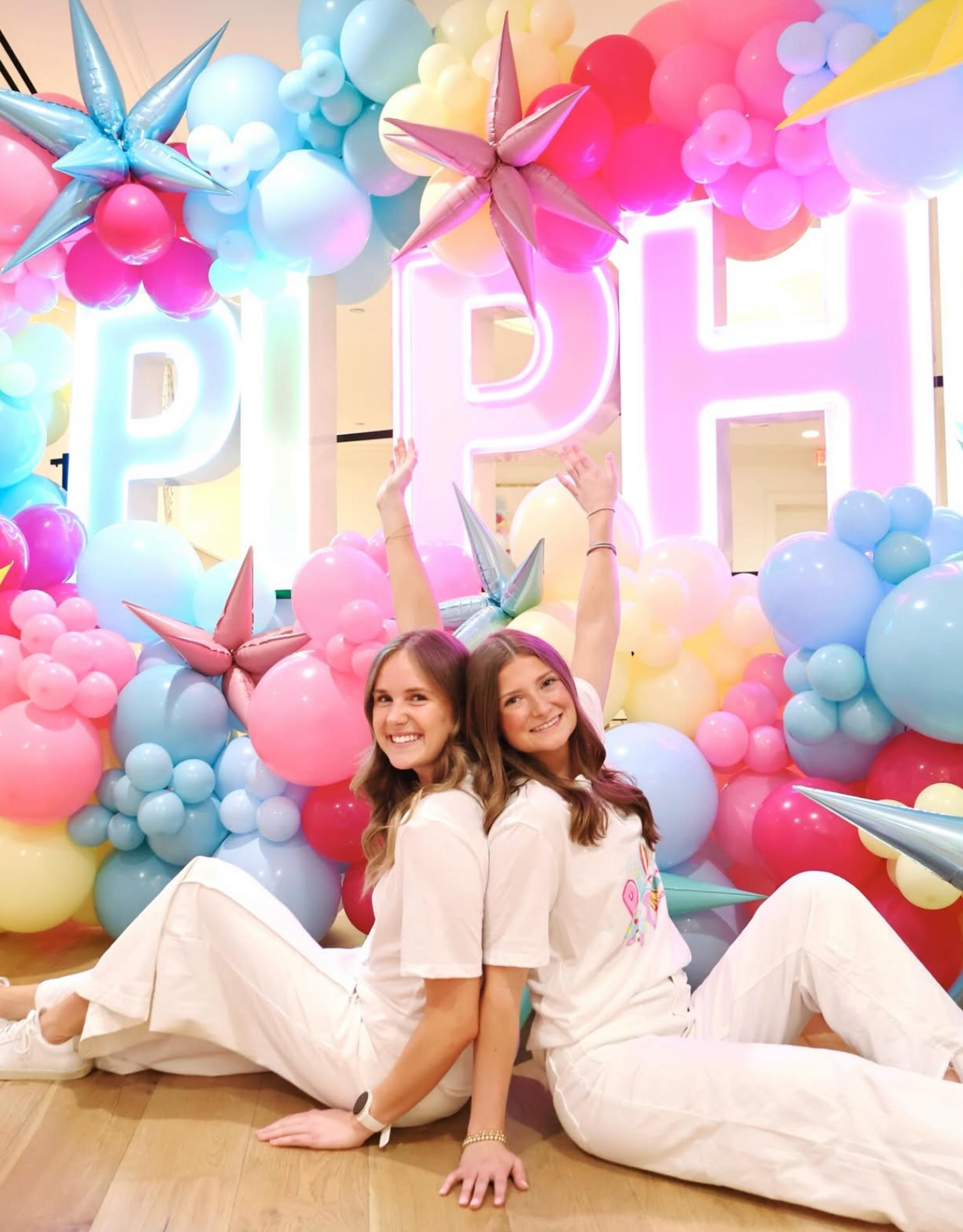 Two individuals sitting indoors in front of giant lights that say Pi Phi