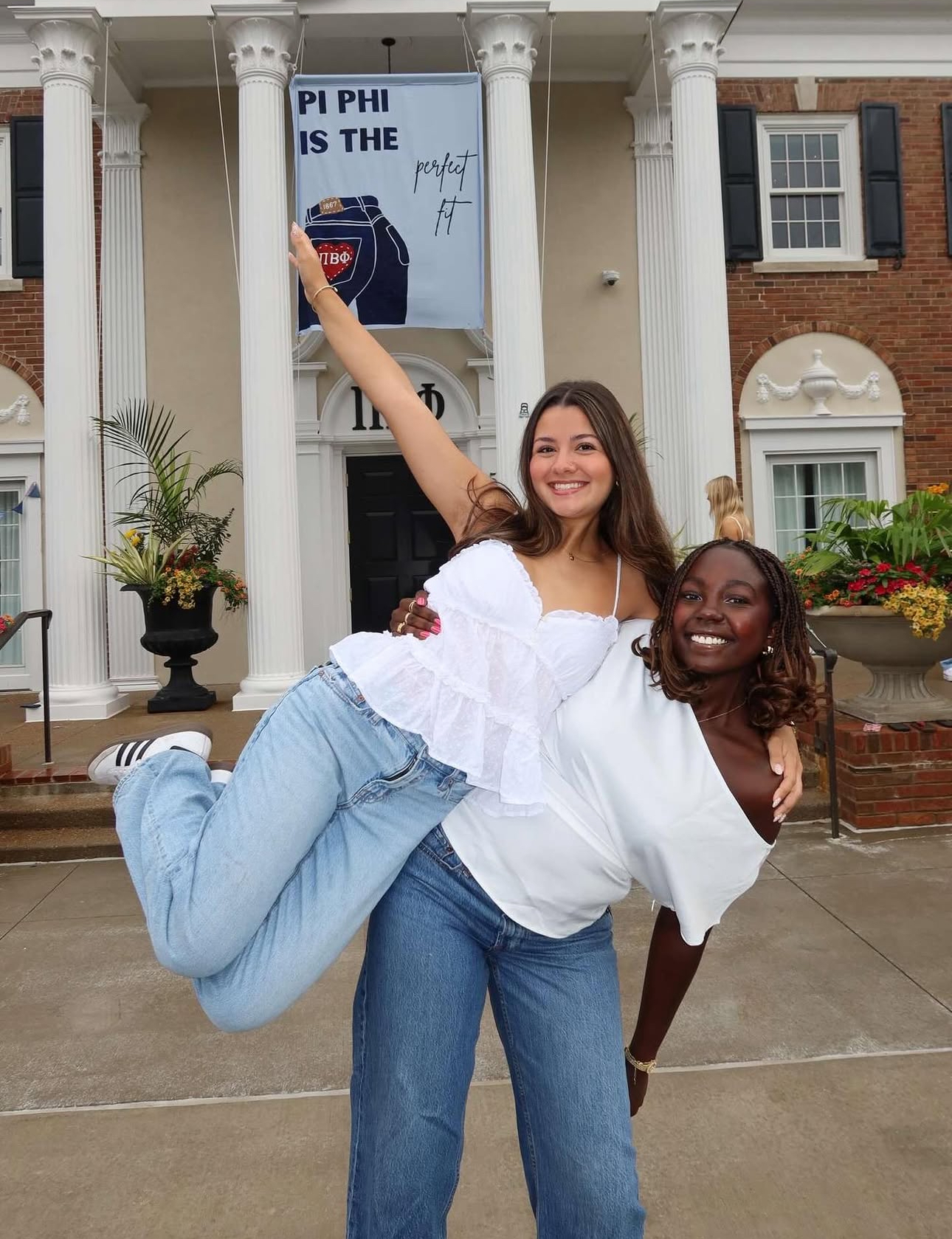 Two members wearing white shirts and blue jeans standing in front of the chapter house. One member is holding the other on their hip
