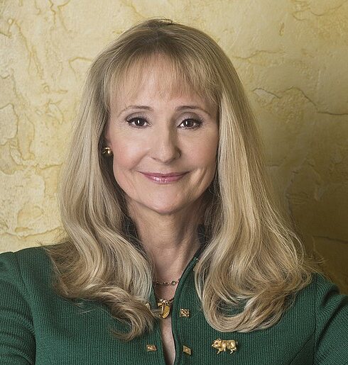 Professional headshot of Nancy Hogshead wearing a green button up jacket with gold buttons in front of a gold background