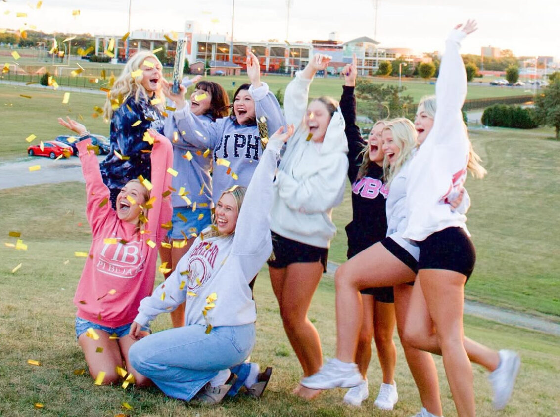 Eight members throwing confetti in the air on a grassy field