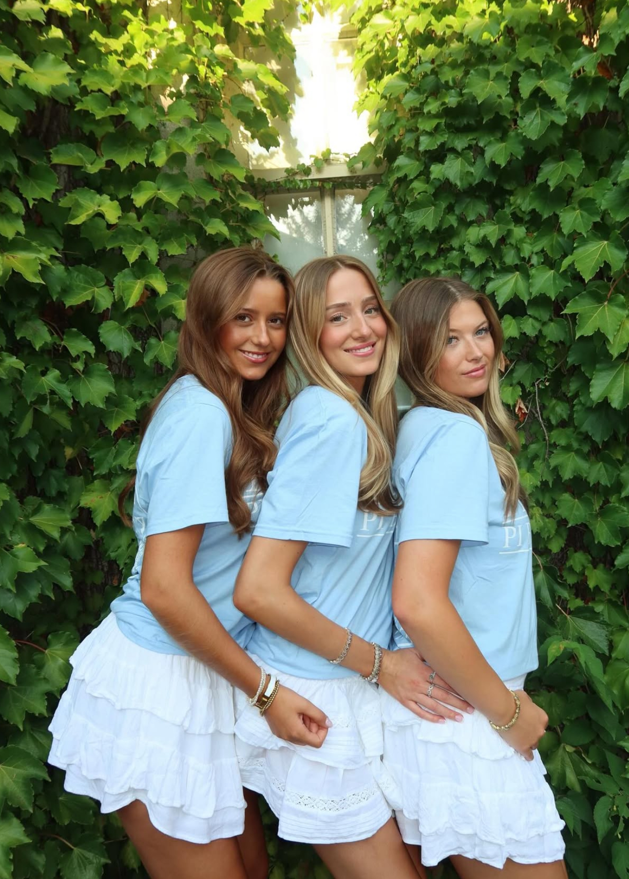 Three members wearing matching blue shirts and white skirts standing in front of a hedge wall