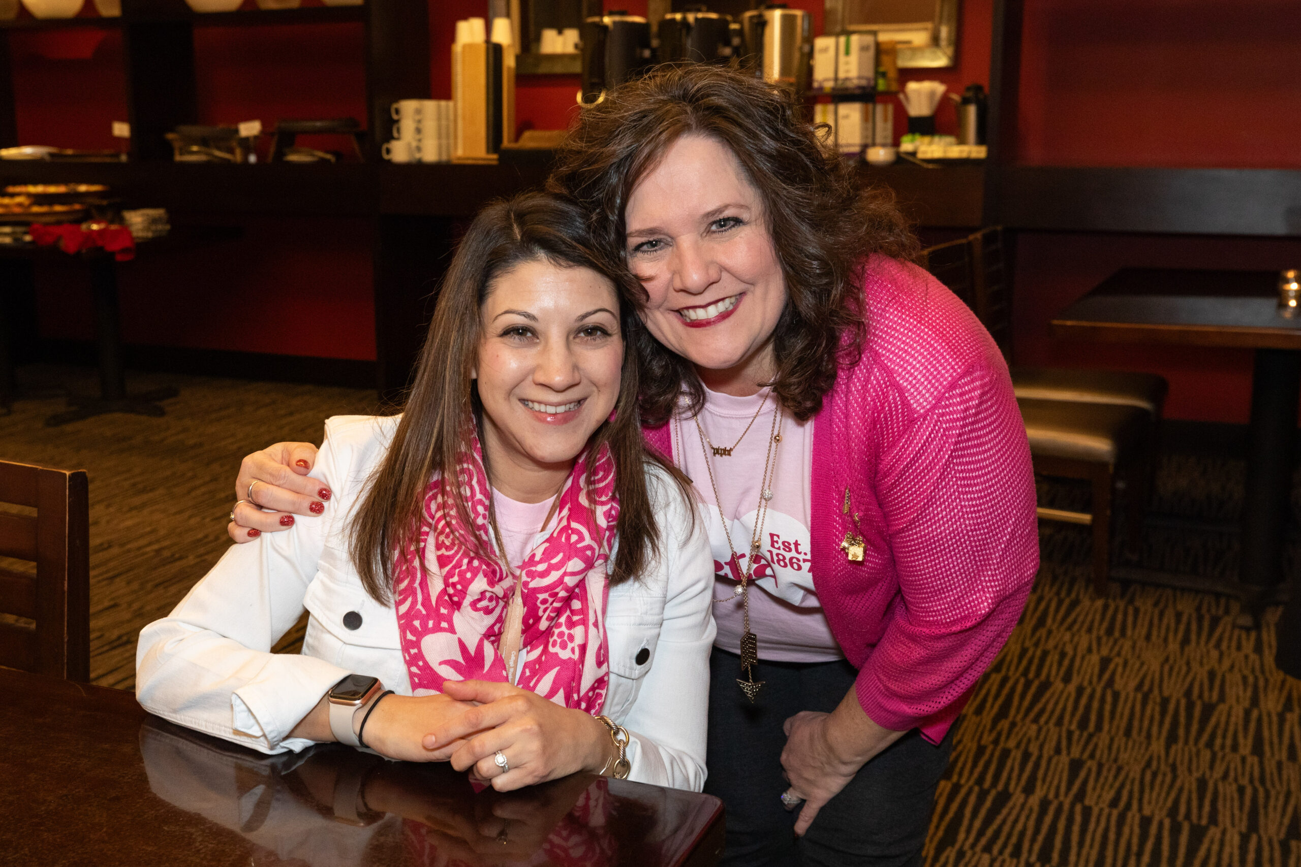 One member sitting with one member leaning over and hugging while both wearing pink and smiling