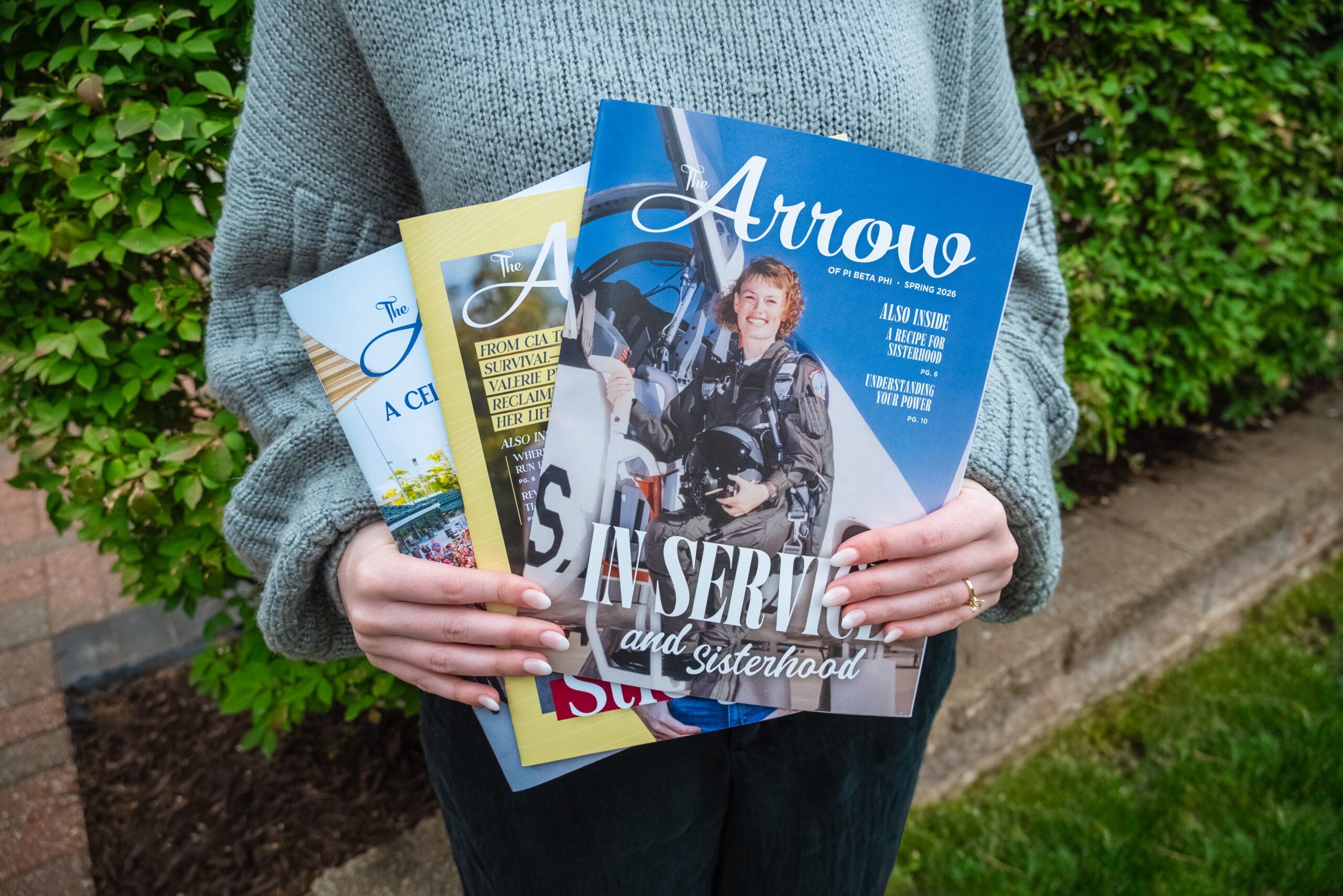 A person standing outdoors holding three copies of The Arrow magazine in front of green bushes