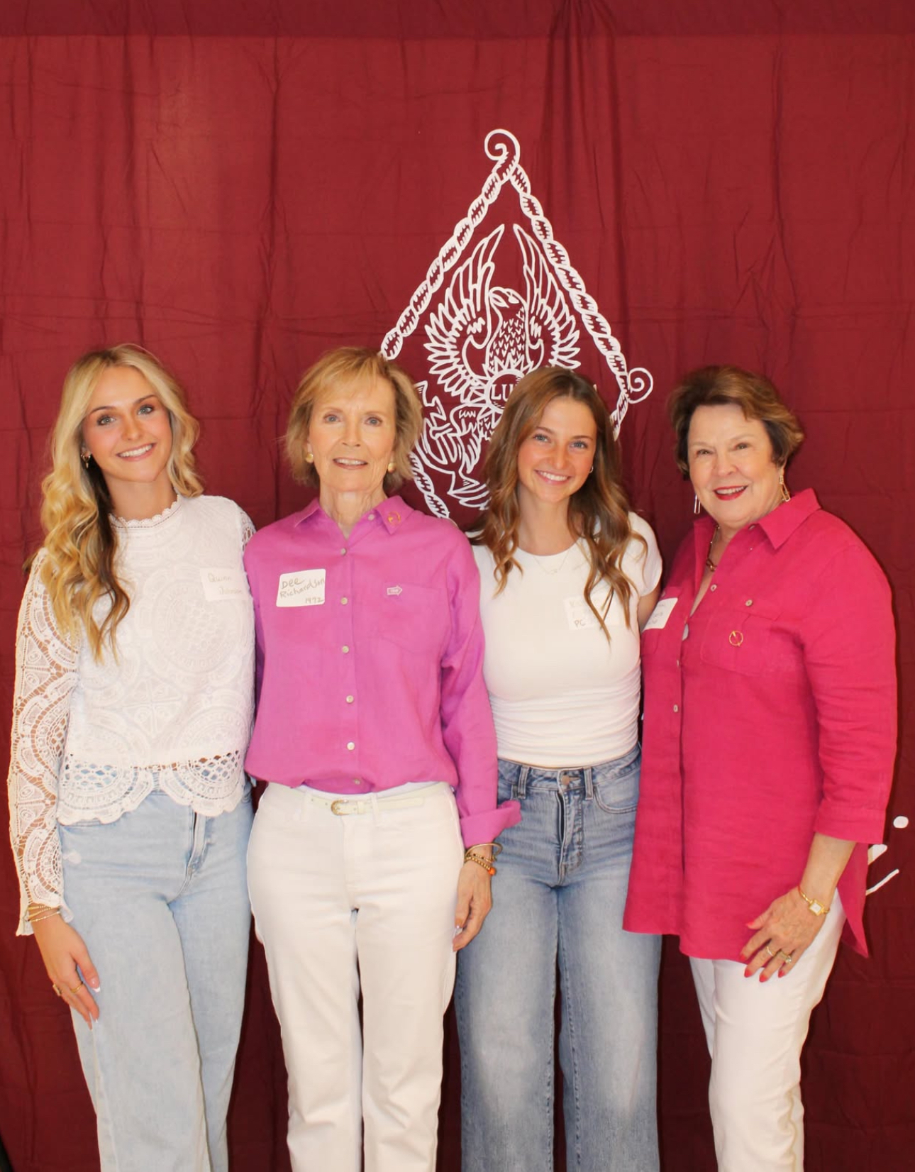 Four alumnae members wearing white, pink and red standing in front of a red wall with a white Pi Beta Phi crest