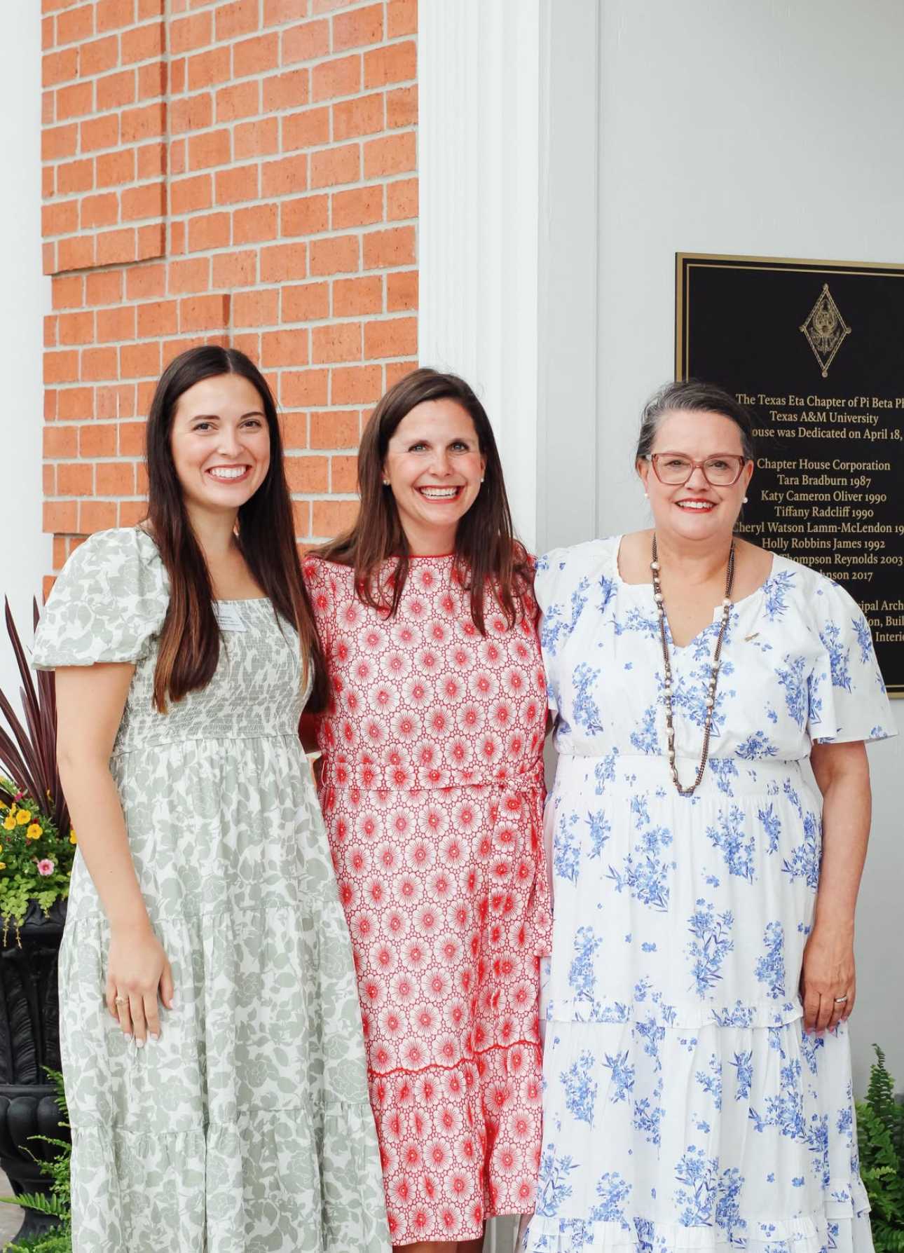 Three members wearing colorful dresses standing outdoors in front of a brick building