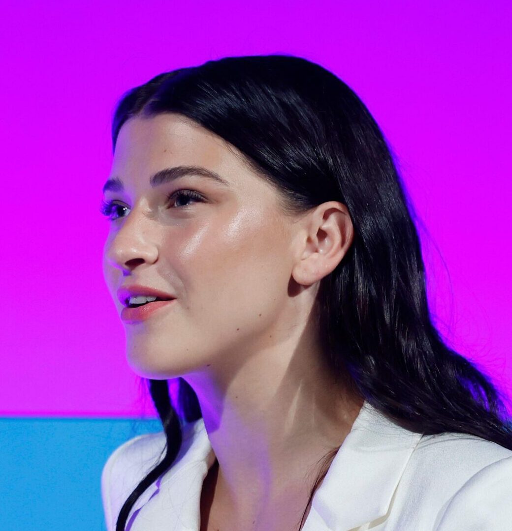 Headshot of Phoebe Gates looking left wearing a white blazer in front of a pink background