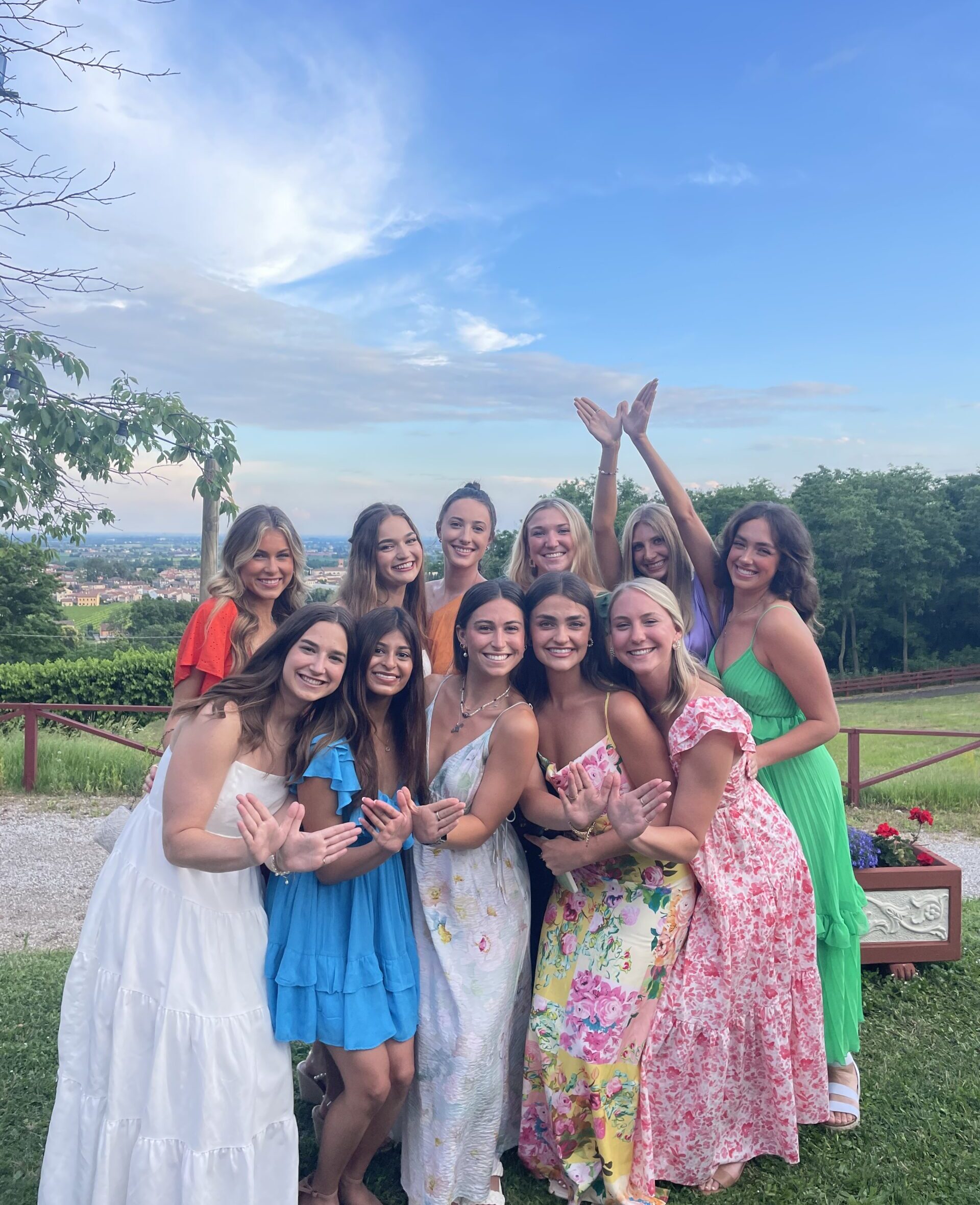 Several members wearing colorful dresses standing outside in front of green trees with one member holding her hands up in angel wings