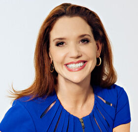 Professional photo of Sally Hogshead sitting with her legs crossed in a blue dress and gold bracelet in front of a white background