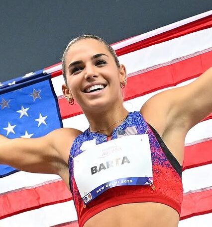 Action shot of Sydney Barta wearing a red and blue athletic top and race bib holding an American flag behind her