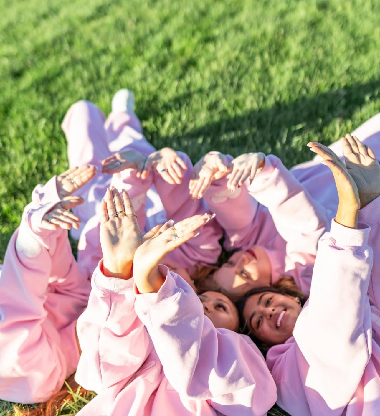 Several members in light pink sweatshirts laying in the grass holding up angel wings with their hands