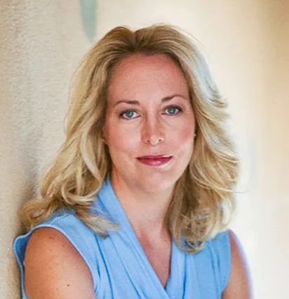 Professional headshot of Valerie Plame Wilson wearing a light blue shirt in front of a beige background
